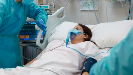 A woman lies in a hospital bed wearing an oxygen mask. She is being spoken to by a healthcare professional wearing full COVID PPE