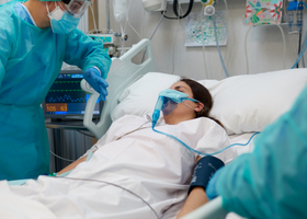A woman lies in a hospital bed wearing an oxygen mask. She is being spoken to by a healthcare professional wearing full COVID PPE