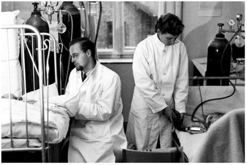 Two doctors ventilating by hand a patient in a hospital bed. They wear white lab coats and the photograph is black and white