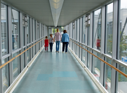 A hospital corridor with glass walls, metal window frames and a pale blue floor with three people in the distance