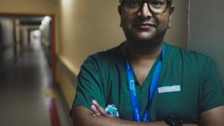 A man wearing green scrubs and an NHS lanyard leans agains the wall in a hospital corridor with his arms crossed.