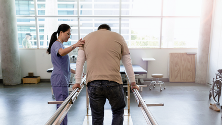 A man walking along a room using wooden bars for support assisted by a woman
