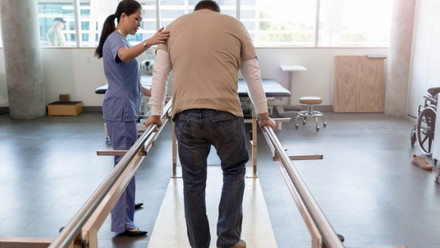 A man walking along a room using wooden bars for support assisted by a woman